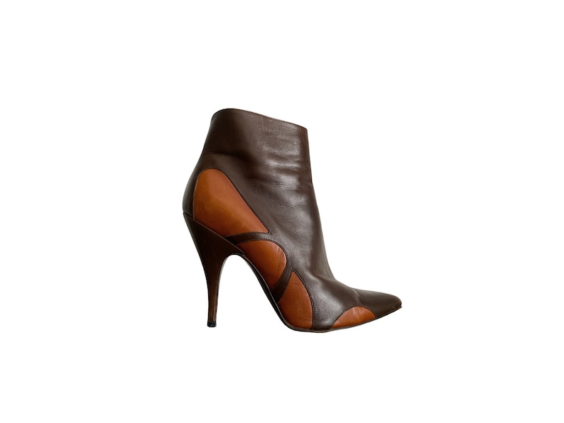 May include: A brown and tan leather ankle boot with a pointed toe and a high stiletto heel. The boot features a unique design with cut-out details and a sleek, modern silhouette. The boot is set against a white background.