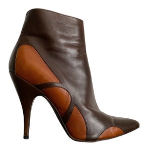 May include: A brown and tan leather ankle boot with a pointed toe and a stiletto heel. The boot features a unique design with tan leather accents and a curved heel. The boot is a fashionable footwear item.