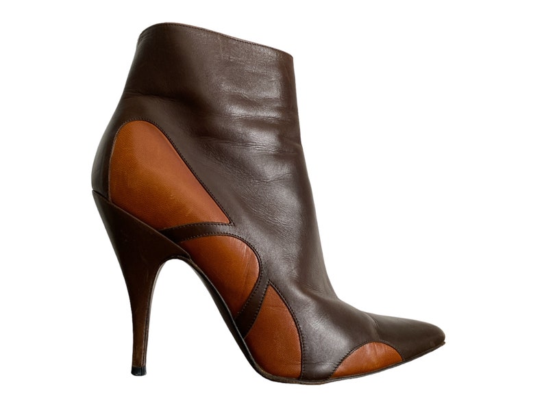 May include: A brown and tan leather ankle boot with a pointed toe and a stiletto heel. The boot features a unique design with tan leather accents and a curved heel. The boot is a fashionable footwear item.