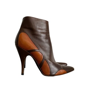 May include: A pair of brown leather ankle boots with a high, stiletto heel. The boots feature a two-tone design, with darker brown leather on the upper and lighter brown accents. The pointed toe and curved design add a stylish touch.