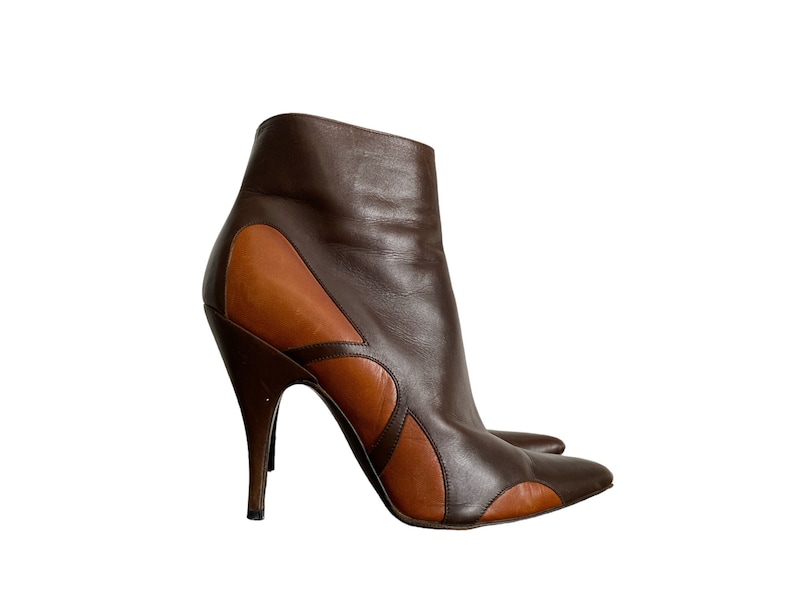 May include: A pair of brown leather ankle boots with a high, stiletto heel. The boots feature a two-tone design, with darker brown leather on the upper and lighter brown accents. The pointed toe and curved design add a stylish touch.