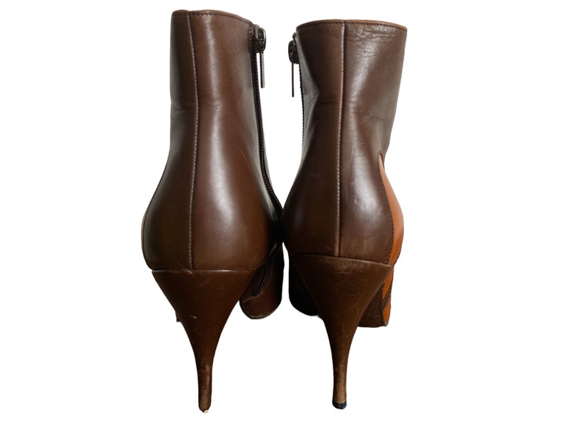 May include: A pair of brown leather ankle boots with stiletto heels. The boots have a zipper closure on the side and a pointed toe. One boot has a patch of tan leather on the side.