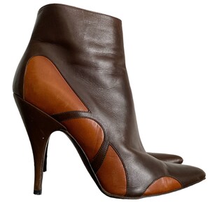 May include: A pair of brown and tan leather ankle boots with a pointed toe and stiletto heel. The boots feature a unique design with curved cutouts and a high, slender heel. The overall height of the boot is approximately 8 inches.