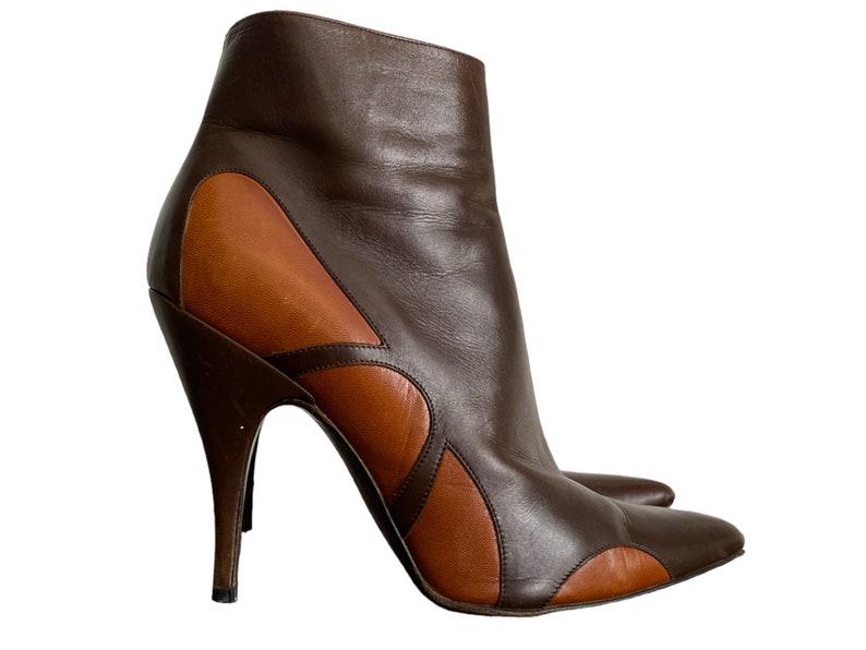 May include: A pair of brown and tan leather ankle boots with a pointed toe and stiletto heel. The boots feature a unique design with curved cutouts and a high, slender heel. The overall height of the boot is approximately 8 inches.