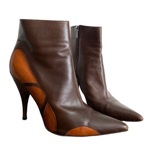 May include: A pair of brown leather ankle boots with a pointed toe and stiletto heel. The boots feature a contrasting tan leather design on the sides and a zipper closure. The heel height is approximately 4 inches.