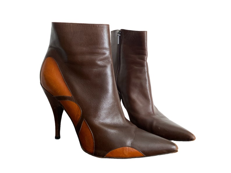 May include: A pair of brown leather ankle boots with a pointed toe and stiletto heel. The boots feature a contrasting tan leather design on the sides and a zipper closure. The heel height is approximately 4 inches.