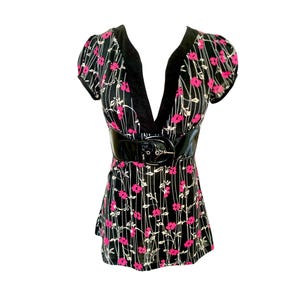 May include: A black short-sleeved top featuring a deep V-neckline with solid black trim and a pattern of small pink flowers and thin white vertical lines. The garment includes a wide, shiny black patent leather belt at the waist.