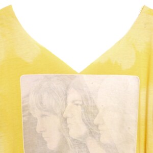 May include: Yellow t-shirt with a faded image of three people with long hair. The image is in a white rectangle.
