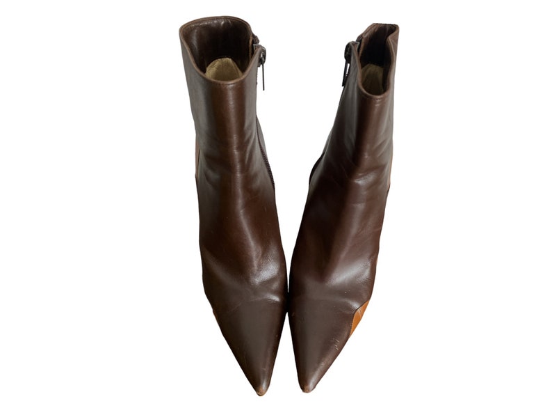 May include: A pair of brown leather ankle boots with pointed toes. The boots have a zipper on the side and a small heel. The leather has a glossy finish. The boots are set against a white background.