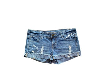 May include: Blue denim shorts with distressed details, including rips and white paint splatters. The shorts have a classic five-pocket design and a button closure. The hem is rolled up.