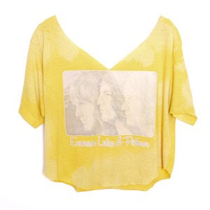 May include: A yellow cropped t-shirt with a faded tie-dye pattern. The shirt features a black and white image of three people with the text "Emerson Lake & Palmer" printed below the image.
