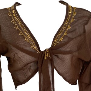 Puede incluir: Un bolero de manga larga en color marr&oacute;n transparente con un escote en V profundo, adornado con lentejuelas y cuentas doradas a lo largo del cuello. La prenda incluye un cierre de lazo frontal hecho de la misma tela marr&oacute;n transl&uacute;cida.