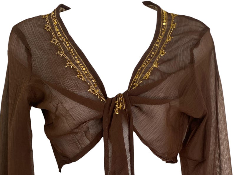 Puede incluir: Un bolero de manga larga en color marr&oacute;n transparente con un escote en V profundo, adornado con lentejuelas y cuentas doradas a lo largo del cuello. La prenda incluye un cierre de lazo frontal hecho de la misma tela marr&oacute;n transl&uacute;cida.