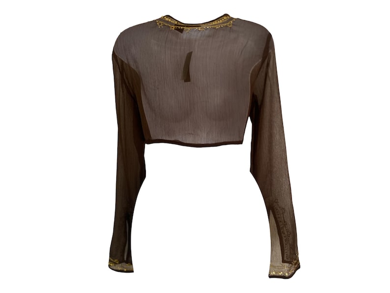 Puede incluir: Un top corto de manga larga en color marr&oacute;n oscuro semitransparente, con un cuello redondo adornado con bordados y detalles en tono dorado. Las mangas largas presentan adornos decorativos en tono dorado a juego en los pu&ntilde;os.