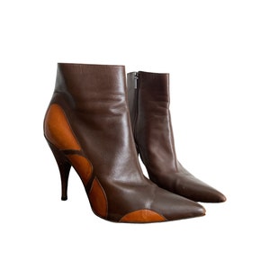 May include: A pair of brown leather ankle boots with a pointed toe and stiletto heel. The boots feature a contrasting tan leather design on the sides and a zipper closure. The heels are approximately 4 inches tall.