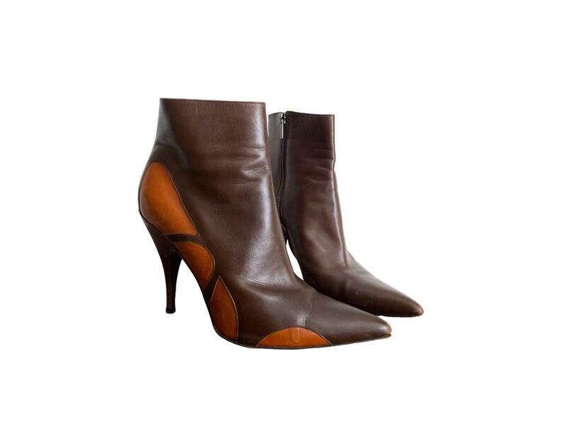 May include: A pair of brown leather ankle boots with a pointed toe and stiletto heel. The boots feature a contrasting tan leather design on the sides and a zipper closure. The heels are approximately 4 inches tall.