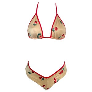 May include: A two-piece lingerie set in beige with a cherry print. The bikini top is a triangle style with red trim and straps. The matching bottoms are low-rise with red trim. The set is designed for comfort and style.