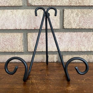May include: A black metal display stand featuring a tripod design with two curved front supports and a central hinged back leg. The frame is constructed from thin, smooth metal rods that terminate in decorative scroll-like curls at the base.
