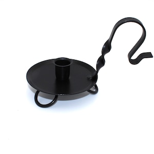 May include: A black metal candlestick holder with a circular base and a curved handle. The holder has a cup for the candle and three small feet. The handle has a twisted design and a hook shape.