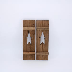 May include: Two rustic wooden shutters with a star cutout design. The shutters are stained a warm brown color and have a distressed appearance. The star cutouts are centered on each shutter, creating a decorative accent. The shutters are designed to be mounted on a wall.