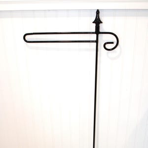 May include: A black metal garden flag stand featuring a vertical pole, a horizontal arm for hanging flags, and a decorative finial at the top. The design includes a curved scroll detail on the side of the arm, providing a stable structure for display.