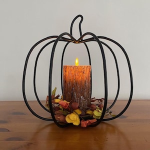 May include: A black metal wire pumpkin frame containing a brown textured pillar candle with a glowing yellow LED flame. The base of the candle is surrounded by a cluster of small artificial autumn-colored leaves and berries in orange and yellow.