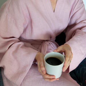 Puede incluir: Una bata de lino rosa claro con cinturón a juego. La bata tiene mangas anchas y se usa mientras se sostiene una taza de café blanca llena de líquido oscuro. La persona está sentada en un sillón estampado.