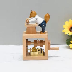 May include: A wooden music box with a squirrel figurine in a white shirt and blue trousers, operating a small machine. The box has a crank, gears, and a gold-coloured mechanism. The squirrel has a brown head and tail.
