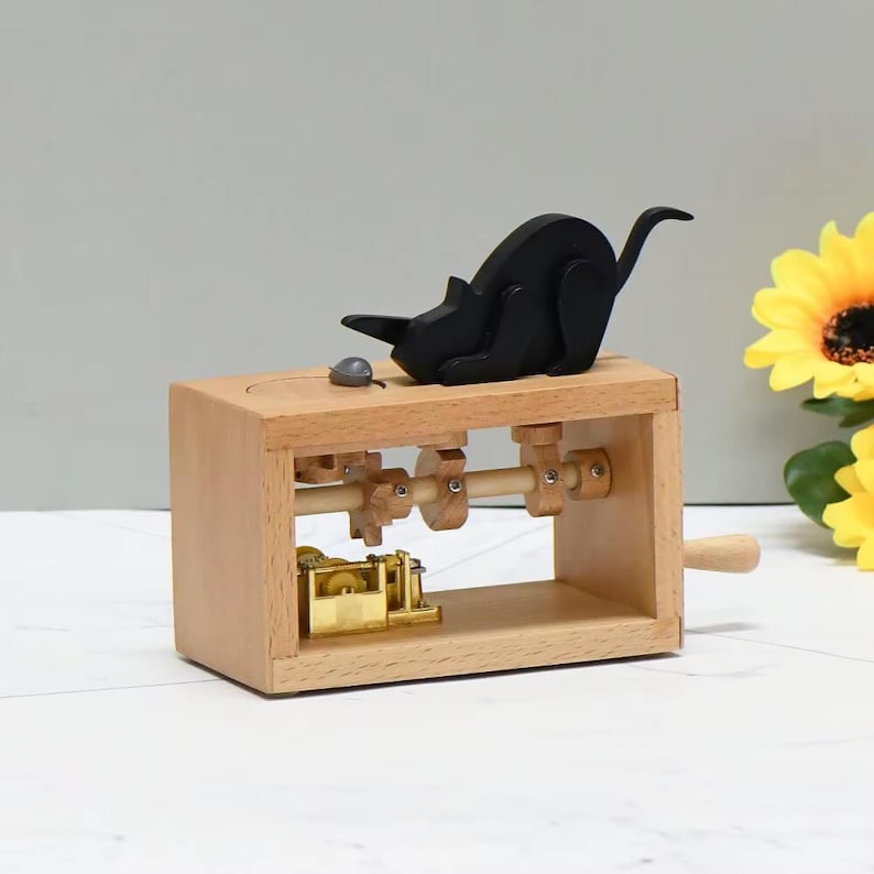 May include: A wooden music box with a black cat figurine and a small grey mouse. The box has visible gears and a hand crank. The cat is made of black wood, and the box is made of light-coloured wood. The box is approximately 15 cm long.