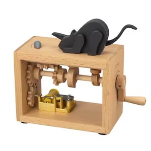 May include: A wooden music box featuring visible gears and a hand crank. A black wooden mouse figure is perched on top. The music box has a gold interior and a small, grey knob on the top.