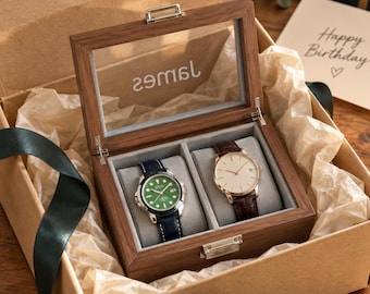 May include: A wooden watch box with a glass lid featuring the name James, containing two wristwatches on grey cushions. One watch has a green face and dark blue strap, while the other has a white face and brown leather strap, all inside a gift box.