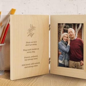 Puede incluir: Un marco de fotos doble plegable de madera clara. El panel izquierdo presenta un diseño floral grabado y el texto: Those we love don't go away. They walk beside us every day. Unseen, unheard, but always near; Still loved, still missed, and always dear. El panel derecho contiene una fotografía de dos personas.