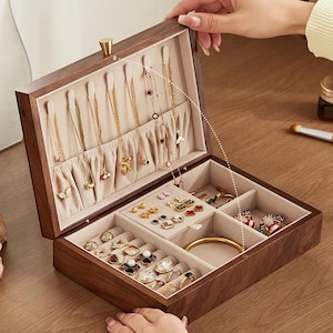 May include: A wooden jewellery box, open to reveal various compartments. The top section displays necklaces, with rings, earrings, and a bracelet in the lower sections. The box is lined with beige fabric and has a gold clasp.