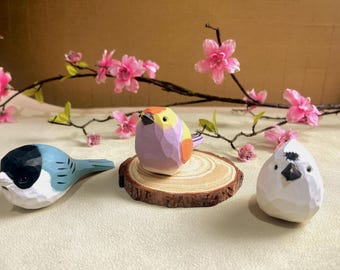 Puede incluir: Tres figuras de pájaros de madera tallada en azul, morado y blanco. El pájaro azul tiene la cabeza negra y detalles blancos. El pájaro morado está sobre una rodaja de madera. Flores de cerezo rosas en el fondo.