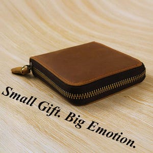 Może przedstawiać: Kwadratowy, brązowy skórzany portfel z zapięciem na zamek błyskawiczny. Portfel leży na jasnej drewnianej powierzchni. Tekst "Small Gift. Big Emotion." jest wydrukowany na drewnianej powierzchni.