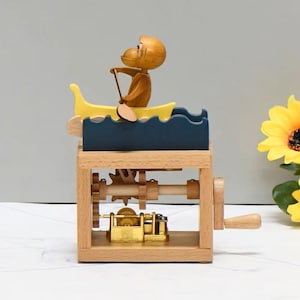 May include: A wooden music box with a monkey in a yellow boat, paddling on a blue wave design. The box has a crank and visible gears. A sunflower is in the background. The box is made of wood.
