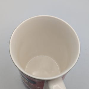 May include: White ceramic mug with a handle. The mug features a red and white design with a cartoon character. The interior of the mug is white. The mug is empty.