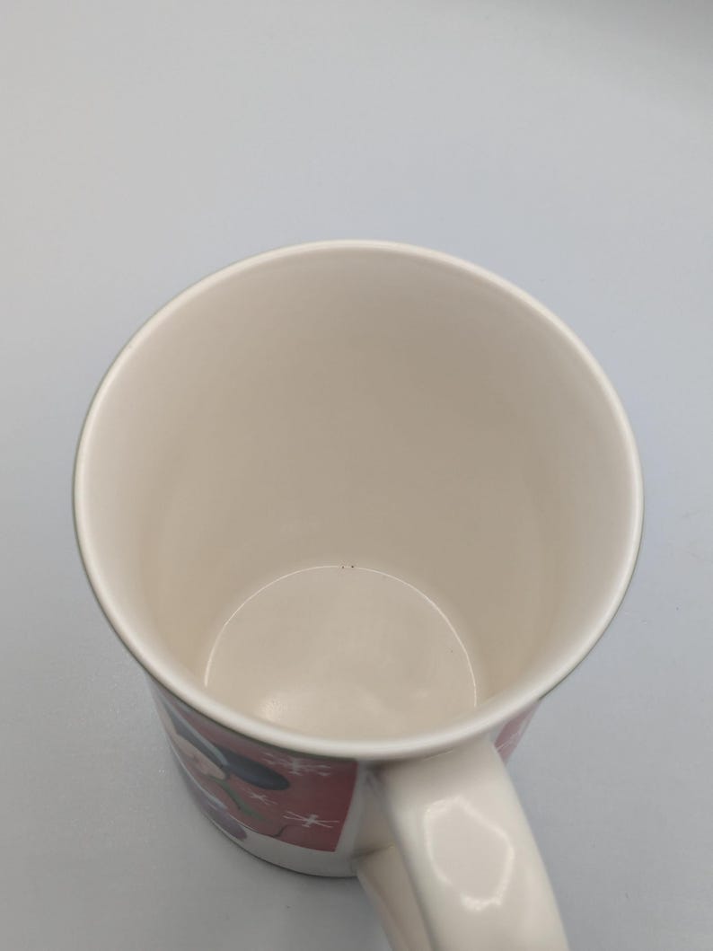 May include: White ceramic mug with a handle. The mug features a red and white design with a cartoon character. The interior of the mug is white. The mug is empty.