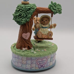 May include: A decorative figurine featuring two teddy bears on a swing set. The bears are dressed in vintage clothing and are set in front of a tree with a blue bird on top. The base of the figurine has the words "Cherished Teddies".