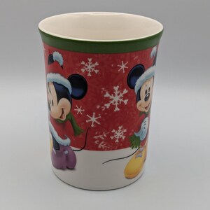 May include: A ceramic mug featuring Mickey Mouse in a winter outfit. The mug has a red background with white snowflakes and a green rim. Mickey is wearing a hat, scarf, and gloves. The mug is approximately 10 cm tall.