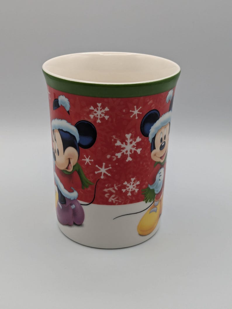 May include: A ceramic mug featuring Mickey Mouse in a winter outfit. The mug has a red background with white snowflakes and a green rim. Mickey is wearing a hat, scarf, and gloves. The mug is approximately 10 cm tall.