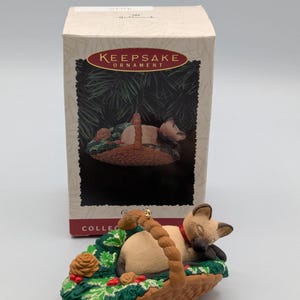 May include: A Hallmark Keepsake Ornament featuring a sleeping cat in a basket. The cat is light brown with a red collar, nestled in a green and brown basket with holly and pinecones. The box behind the ornament reads "Keepsake Ornament".