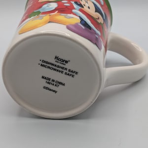May include: White ceramic mug featuring Mickey and Minnie Mouse in festive holiday attire. The mug has a green rim and a white handle. The bottom of the mug has text that reads "Dishwasher Safe" and "Microwave Safe".