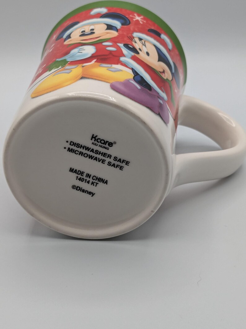 May include: White ceramic mug featuring Mickey and Minnie Mouse in festive holiday attire. The mug has a green rim and a white handle. The bottom of the mug has text that reads "Dishwasher Safe" and "Microwave Safe".