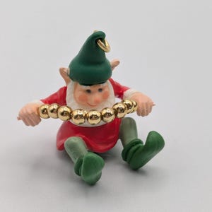 May include: A small figurine of a gnome wearing a green hat, red tunic, and green pants, holding a horizontal string of shiny gold-colored spherical beads in its hands. The gnome has a white beard and a gold-colored metal loop attached to the hat.