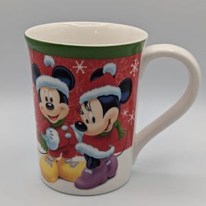 May include: A white ceramic mug featuring Mickey and Minnie Mouse in winter attire. The mug has a red background with snowflakes and a green rim. Mickey Mouse wears a red coat, yellow shoes, and a winter hat. Minnie Mouse wears a red coat, purple shoes, and a winter hat.