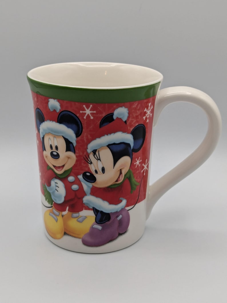 May include: A white ceramic mug featuring Mickey and Minnie Mouse in winter attire. The mug has a red background with snowflakes and a green rim. Mickey Mouse wears a red coat, yellow shoes, and a winter hat. Minnie Mouse wears a red coat, purple shoes, and a winter hat.