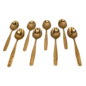 May include: Eight gold-colored spoons with ornate, textured handles and rounded bowls are arranged on a white background. The spoons are evenly spaced and angled, showcasing their decorative design. The spoons appear to be made of metal.