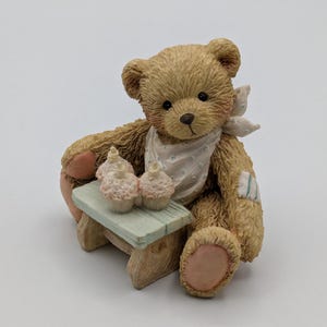 May include: A brown teddy bear figurine with a white bib and a light blue bandage. The bear is seated next to a small, light blue table holding three pink cupcakes. The figurine has a charming, whimsical design.