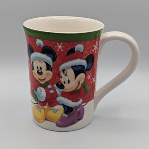 May include: A white ceramic mug featuring Mickey and Minnie Mouse in winter attire. The mug has a red background with snowflakes and a green rim. Mickey and Minnie are wearing red coats and hats.